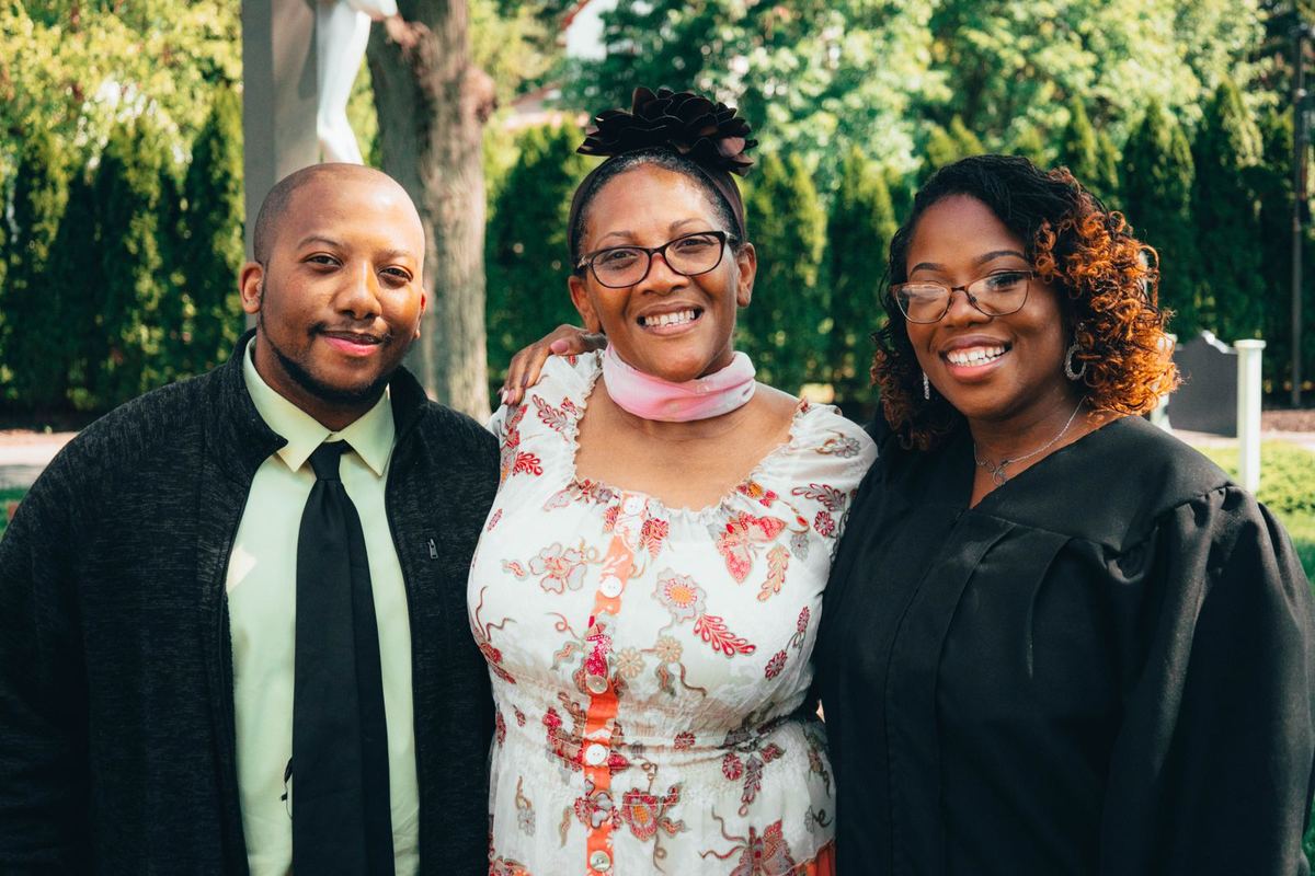 A graduate in her gown beaming beside her mother and brother in golden evening light