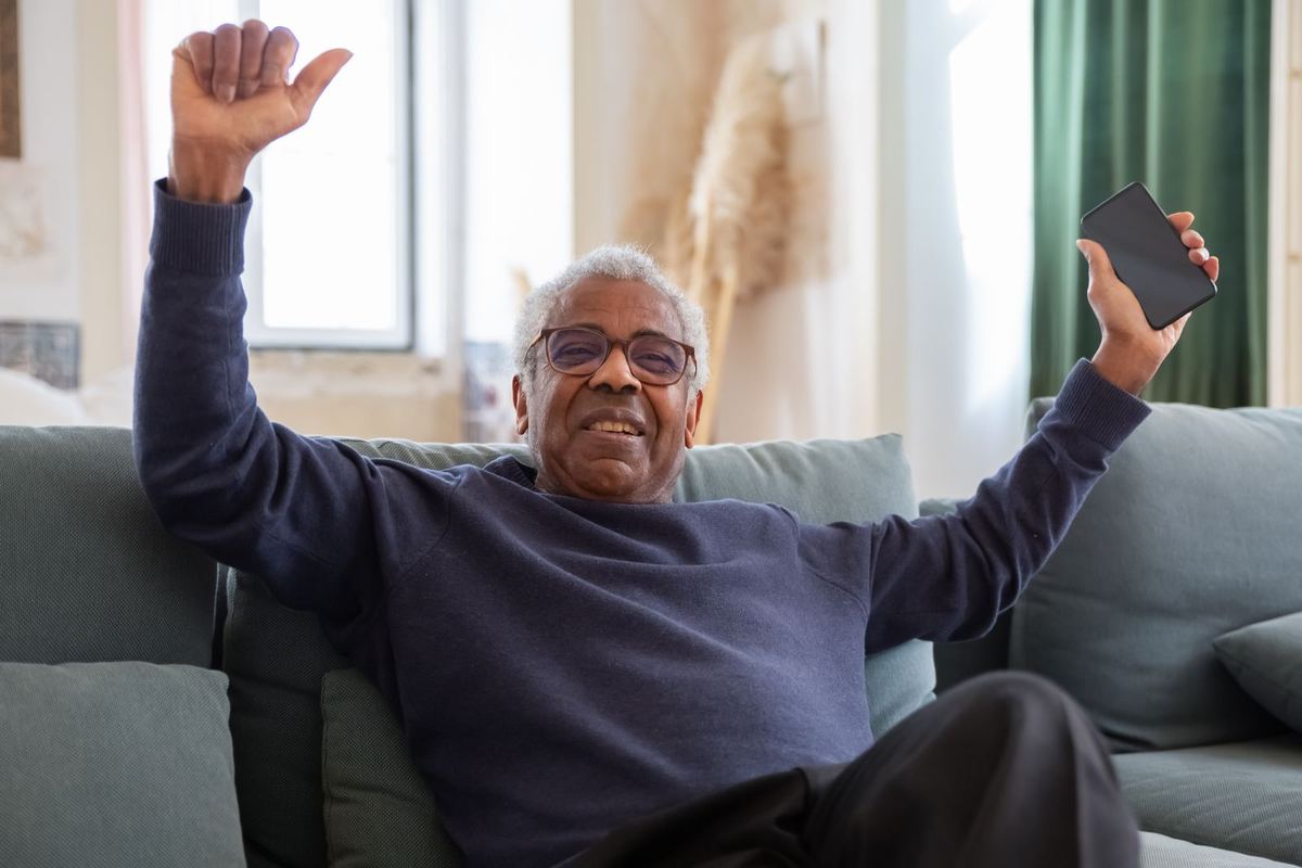 A silver-haired man throwing both arms up in delight, phone in hand, in a bright living room