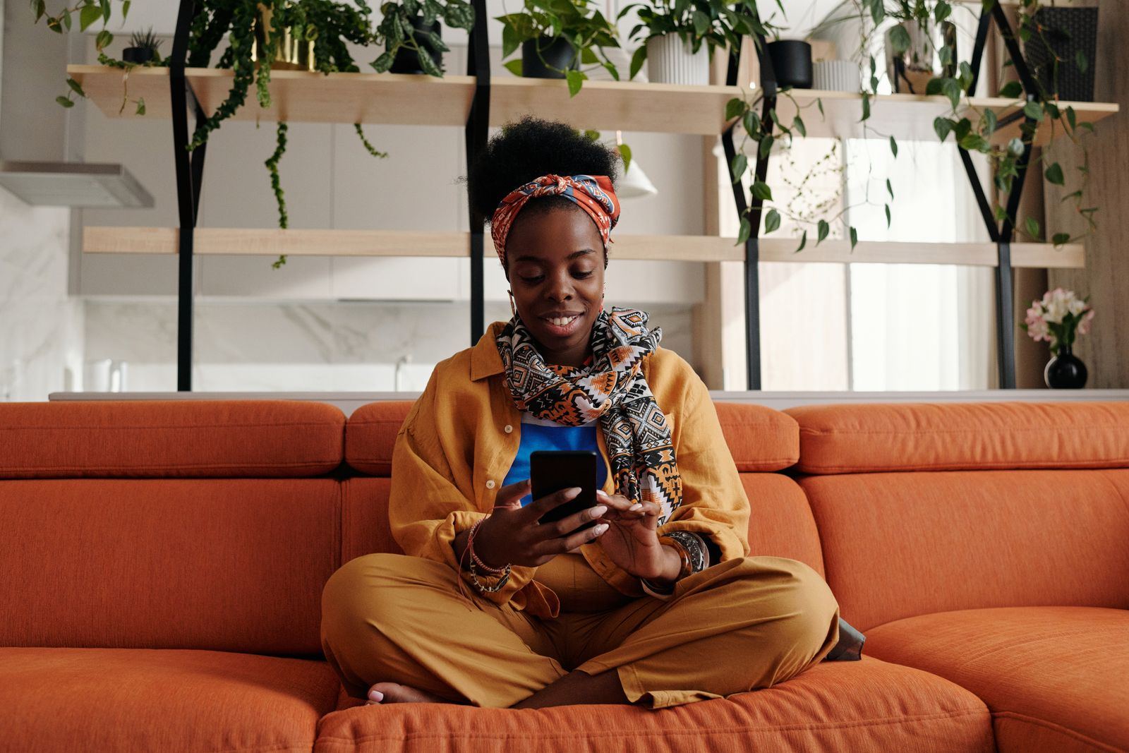 A woman in an Ankara headband smiling at her phone on a terracotta sofa